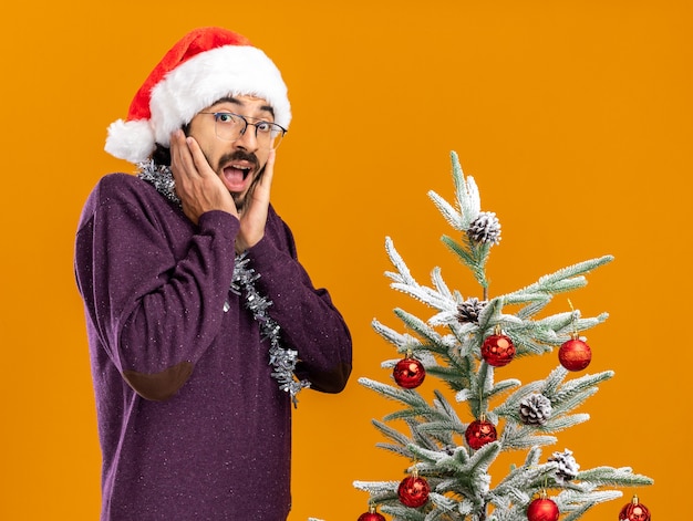 Chico guapo joven emocionado de pie cerca del árbol de navidad con sombrero de navidad con guirnalda en el cuello poniendo las manos en las mejillas aisladas en la pared naranja