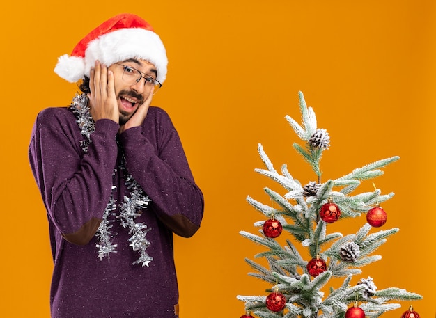 Chico guapo joven emocionado de pie cerca del árbol de Navidad con sombrero de Navidad con guirnalda en el cuello poniendo las manos en las mejillas aisladas sobre fondo naranja