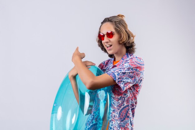 Chico guapo joven con gafas de sol rojas con anillo inflable sonriendo alegremente apuntando a la espalda detrás de pie