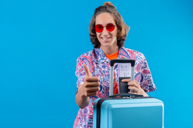 Foto gratuita chico guapo joven con gafas de sol rojas con maleta de viaje con billetes de avión mostrando los pulgares para arriba sonriendo feliz y positivo de pie alegremente sobre fondo azul