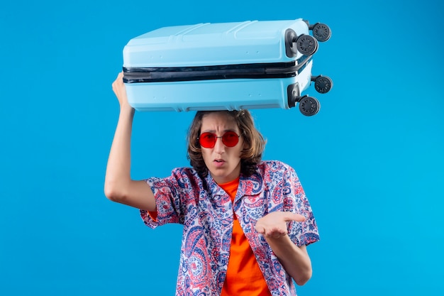 Foto gratuita chico guapo joven con gafas de sol rojas con maleta de viaje despistado y confundido sin tener respuesta extendiendo los brazos de pie