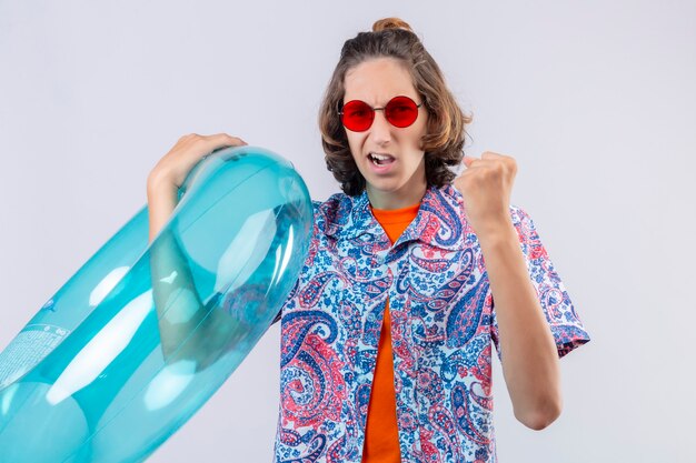 Foto gratuita chico guapo joven con gafas de sol rojas sosteniendo el anillo inflable levantando el puño con expresión enojada de pie sobre fondo blanco.