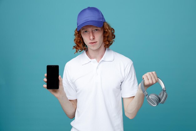 Chico guapo joven en gorra sosteniendo auriculares con auriculares de teléfono aislado sobre fondo azul.