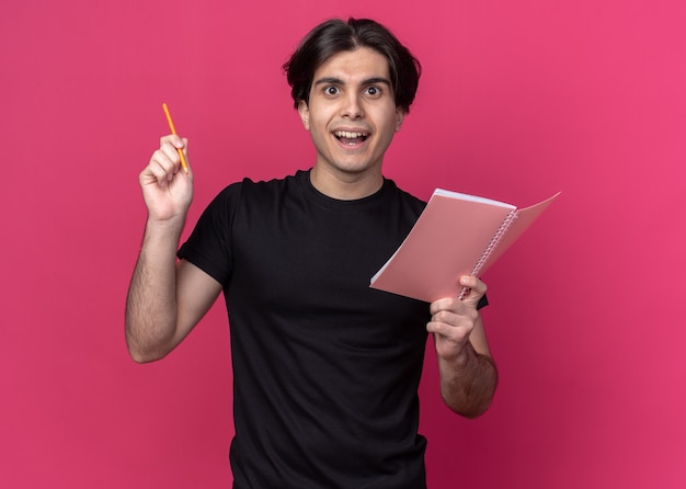 Chico guapo joven impresionado con camiseta negra con cuaderno con lápiz - aislado en la pared rosa