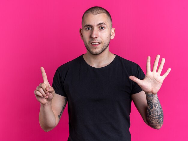 Chico guapo joven impresionado con camiseta negra que muestra diferentes números aislados sobre fondo rosa