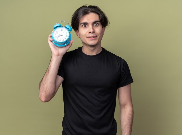 Chico guapo joven impresionado con camiseta negra con reloj despertador alrededor de la cara aislada en la pared verde oliva