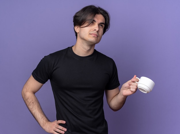Chico guapo joven impresionado con camiseta negra sosteniendo una taza de café poniendo la mano en la cadera aislada en la pared púrpura