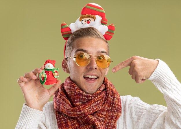 Chico guapo joven impresionado con diadema y bufanda de santa claus mirando a la cámara sosteniendo y apuntando al muñeco de nieve adorno navideño aislado sobre fondo verde oliva