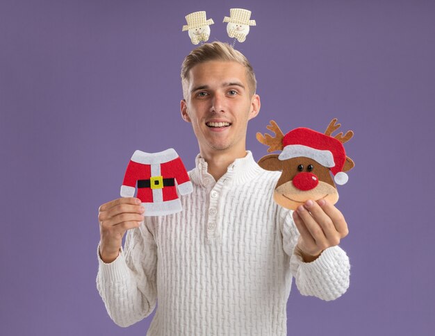 Chico guapo joven impresionado con diadema de muñeco de nieve sosteniendo y estirando adornos de papel de Navidad hacia aislados en la pared púrpura