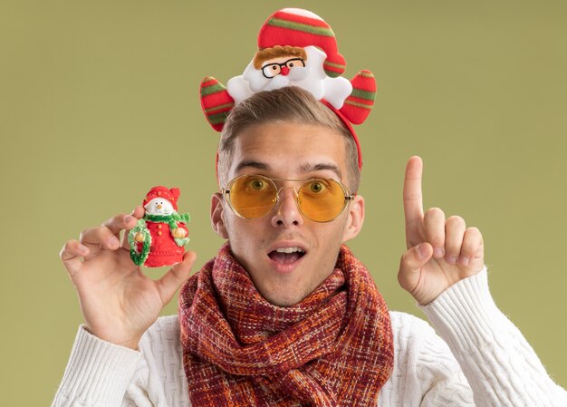 Chico guapo joven impresionado con diadema de santa claus y bufanda sosteniendo muñeco de nieve adorno de navidad apuntando hacia arriba aislado en la pared verde oliva