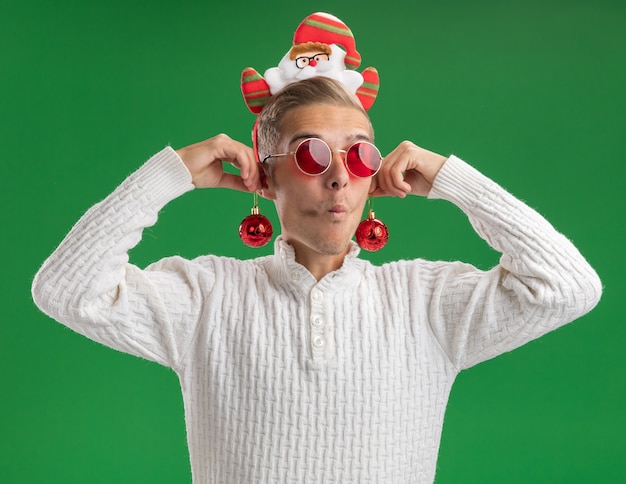 Foto gratuita chico guapo joven impresionado con diadema de santa claus con gafas colgando adornos navideños en las orejas mirando al lado con los labios fruncidos aislados en la pared verde