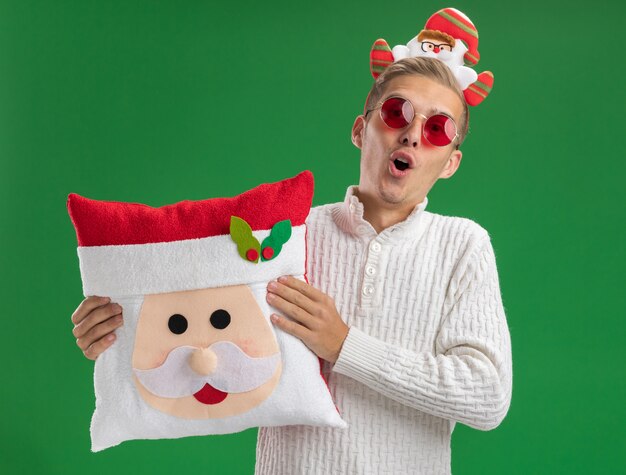 Foto gratuita chico guapo joven impresionado con diadema de santa claus con gafas sosteniendo almohada de santa claus aislado en la pared verde