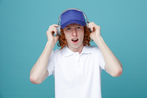 Chico guapo joven impresionado en gorra usando auriculares aislados sobre fondo azul