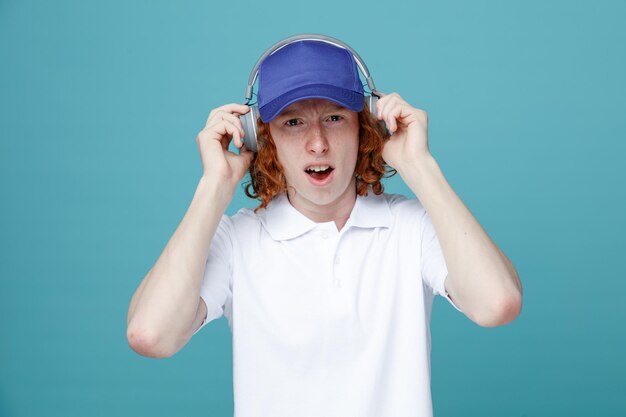 Chico guapo joven impresionado en gorra usando auriculares aislados sobre fondo azul