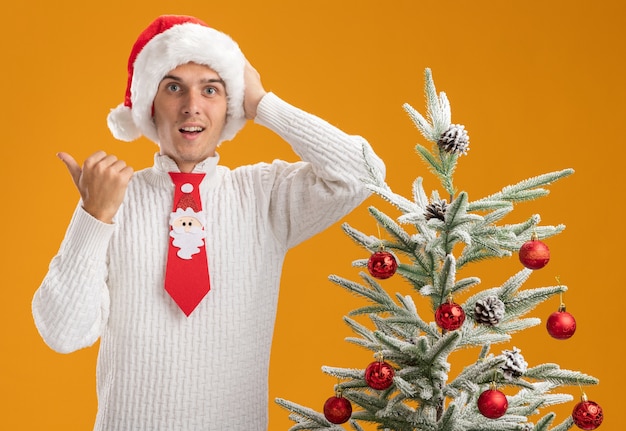 Chico guapo joven impresionado con sombrero de navidad y corbata de santa claus de pie cerca del árbol de navidad decorado poniendo la mano en la cabeza apuntando al lado aislado en la pared naranja