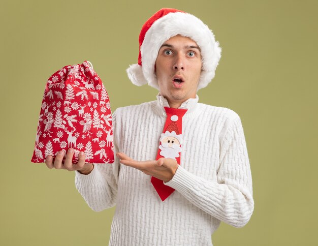 Chico guapo joven impresionado con sombrero de navidad y corbata de santa claus sosteniendo y apuntando con la mano al saco de navidad aislado en la pared verde oliva