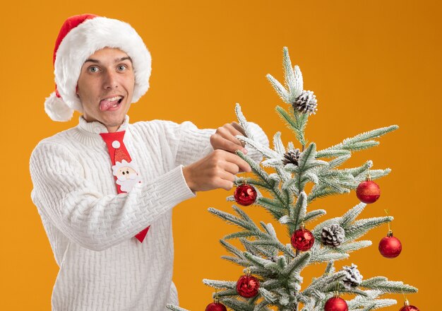 Chico guapo joven juguetón con sombrero de navidad y corbata de santa de pie cerca del árbol de navidad adornándolo con adornos de bolas de navidad