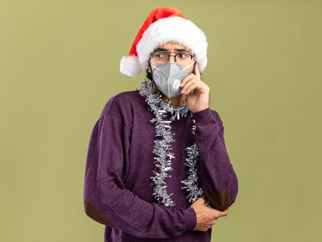 Chico guapo joven de lado mirando preocupado con sombrero de navidad y máscara médica con guirnalda en el cuello poniendo la mano en la mejilla aislada en la pared verde oliva