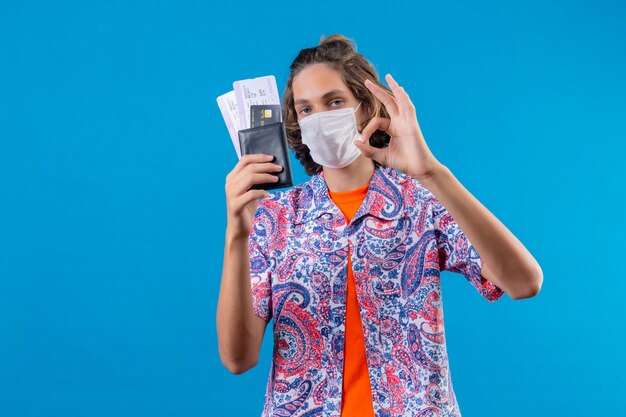 Foto gratuita chico guapo joven con máscara protectora facial sosteniendo boletos de avión haciendo bien firmar mirando con expresión de confianza de pie sobre fondo azul