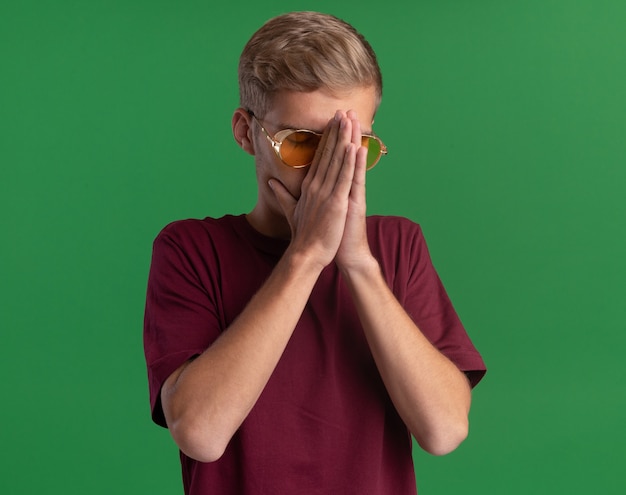 Chico guapo joven con los ojos cerrados con camisa roja y gafas agarró la nariz con las manos aisladas en la pared verde