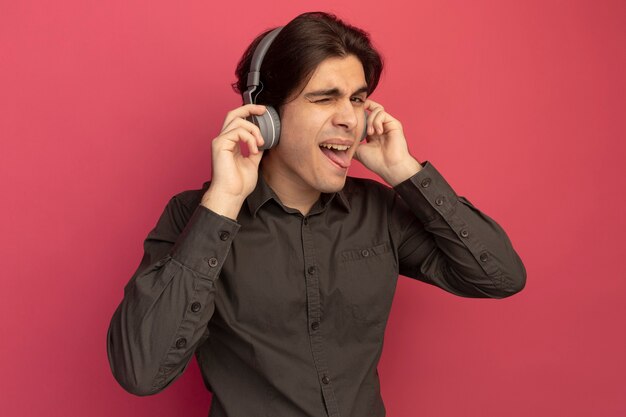 Chico guapo joven parpadeó con camiseta negra con auriculares mostrando la lengua aislada en la pared rosa