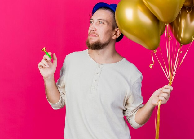 Chico guapo joven partido eslavo con sombrero de fiesta sosteniendo globos y soplador de fiesta mirando al lado aislado en la pared rosa