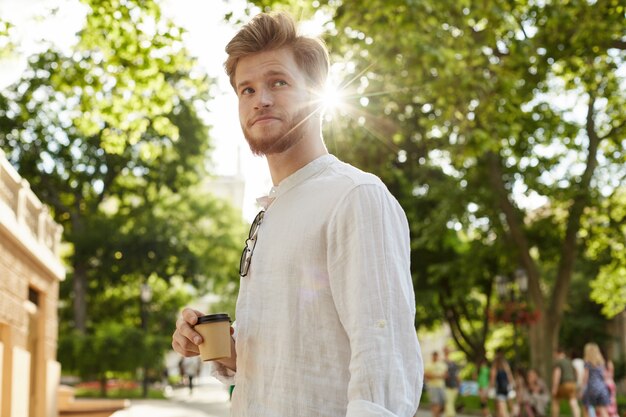 Chico guapo joven con pelo de jengibre y barba en camisa blanca en el parque