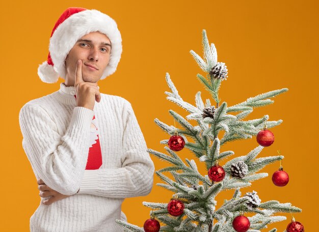 Chico guapo joven pensativo con sombrero de navidad y corbata de santa claus de pie cerca del árbol de navidad decorado poniendo la mano en la barbilla mirando a cámara aislada sobre fondo naranja