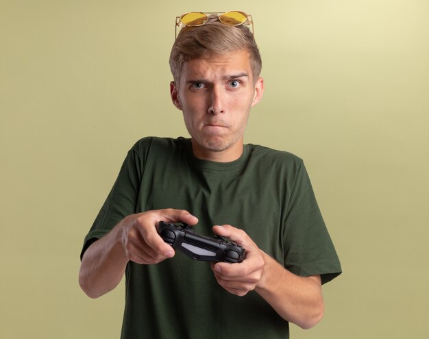 Chico guapo joven preocupado con camisa verde con gafas en la cabeza jugando en el joystick del controlador del juego aislado en la pared verde oliva