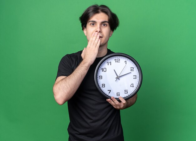 Chico guapo joven preocupado con camiseta negra sosteniendo el reloj de pared con la boca cubierta con la mano aislada en la pared verde con espacio de copia