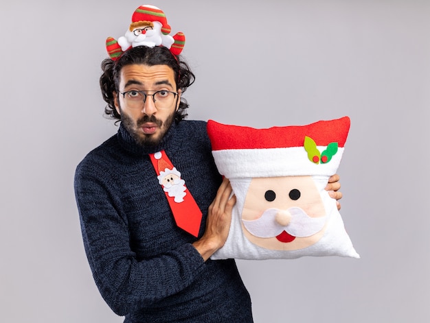 Chico guapo joven preocupado vistiendo corbata de Navidad con aro de pelo sosteniendo almohada de Navidad aislado en la pared blanca