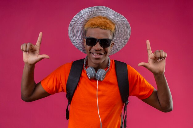 Chico guapo joven con sombrero de verano con cámara y auriculares feliz y positivo apuntando con el dedo hacia arriba de pie sobre la pared rosa