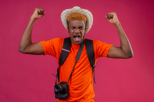 Chico guapo joven con sombrero de verano con cámara con expresión enojada levantando puños sobre pared rosa