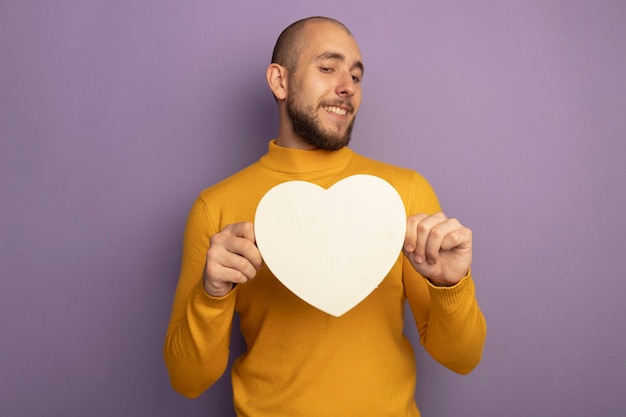 Chico guapo joven sonriente con caja en forma de corazón aislada en púrpura