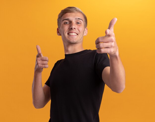 Chico guapo joven sonriente con camisa negra que le muestra gesto aislado en la pared amarilla