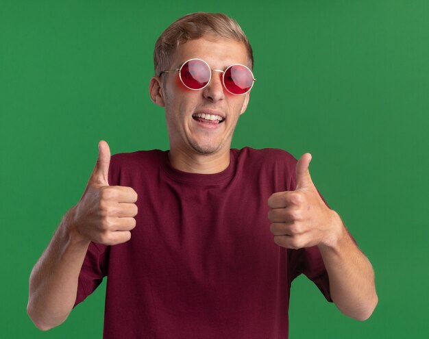 Chico guapo joven sonriente con camisa roja y gafas mostrando los pulgares para arriba aislado en la pared verde