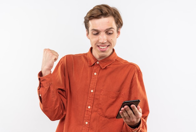 Chico guapo joven sonriente con camisa roja sosteniendo y mirando el teléfono mostrando sí gesto aislado en la pared blanca