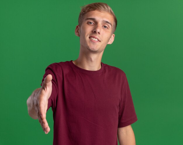 Chico guapo joven sonriente con camisa roja tendiendo la mano a la cámara aislada en la pared verde