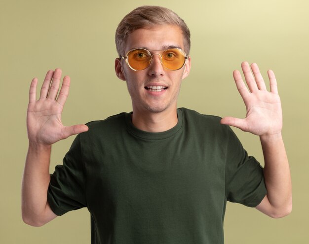 Chico guapo joven sonriente con camisa verde con gafas levantando las manos aisladas en la pared verde oliva