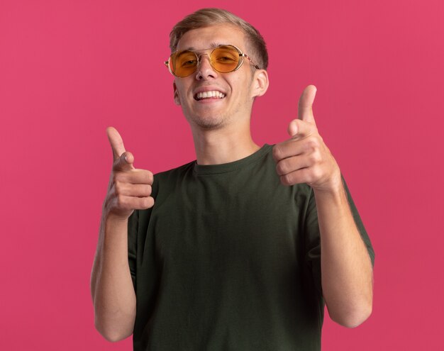 Chico guapo joven sonriente con camisa verde y gafas mostrando gesto aislado en la pared rosa