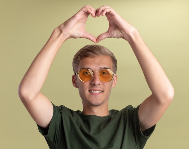 Chico guapo joven sonriente con camisa verde con gafas mostrando gesto de corazón aislado en la pared verde oliva