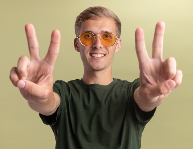 Chico guapo joven sonriente con camisa verde con gafas mostrando gesto de paz aislado en la pared verde oliva