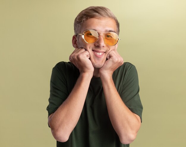 Chico guapo joven sonriente con camisa verde con gafas poniendo puños en las mejillas aisladas en la pared verde oliva