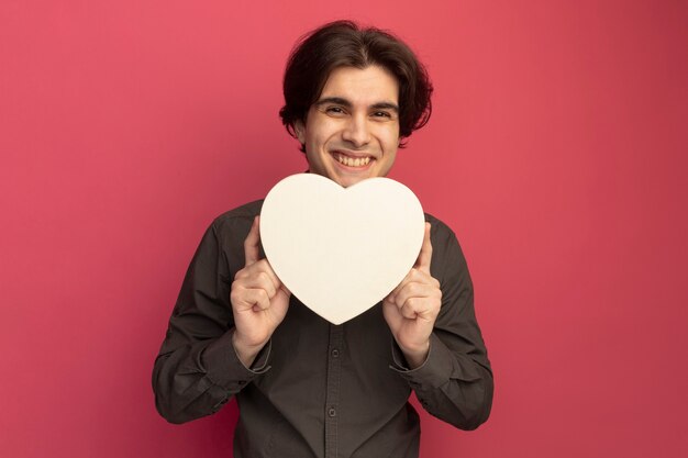 Foto gratuita chico guapo joven sonriente con camiseta negra con caja en forma de corazón aislada en la pared rosa