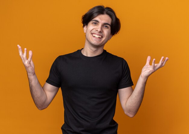 Chico guapo joven sonriente con camiseta negra extendiendo las manos aisladas en la pared naranja