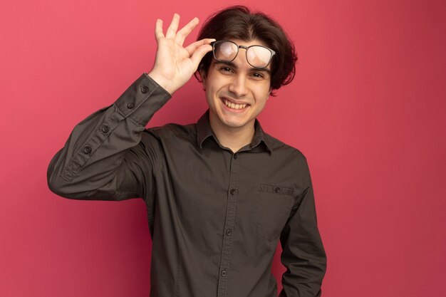 Chico guapo joven sonriente con camiseta negra y gafas aisladas en la pared rosa