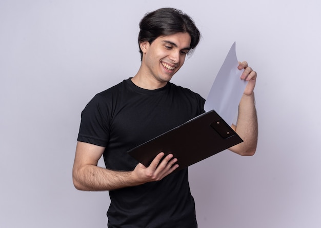 Chico guapo joven sonriente con camiseta negra hojeando el portapapeles aislado en la pared blanca