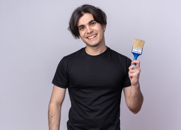 Chico guapo joven sonriente con camiseta negra con pincel aislado en la pared blanca