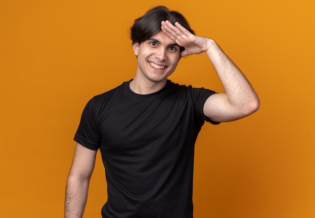 Foto gratuita chico guapo joven sonriente con camiseta negra que muestra gesto de saludo aislado en la pared naranja