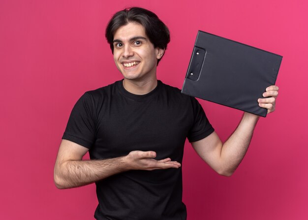 Chico guapo joven sonriente con camiseta negra sosteniendo y apunta al portapapeles aislado en la pared rosa